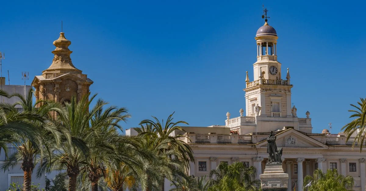Ayuntamiento de Cádiz con arquitectura icónica y estatua, enmarcado por palmeras bajo un cielo azul claro.