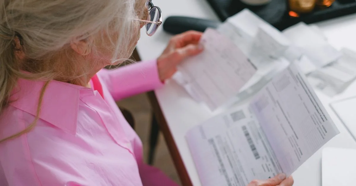 Una mujer anciana con gafas sostiene y lee documentos importantes en una mesa en el interior.