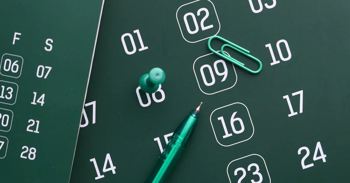 Primer plano de un calendario verde con bolígrafo, chincheta y clip de papel para planificación y organización.