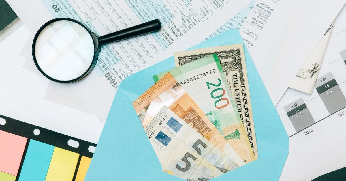 Primer plano de documentos fiscales y moneda mixta en un sobre azul con una lupa, perfecto para conceptos de análisis financiero.